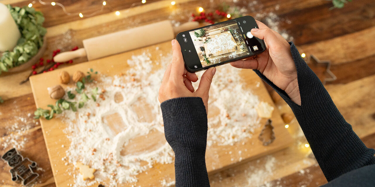 Môžete vidieť veľký drevený valček posypaný múkou a zdobený na Vianoce. V popredí môžete vidieť dve ruky, ktoré držia smartphone vo vodorovnej polohe a fotografujú valec.