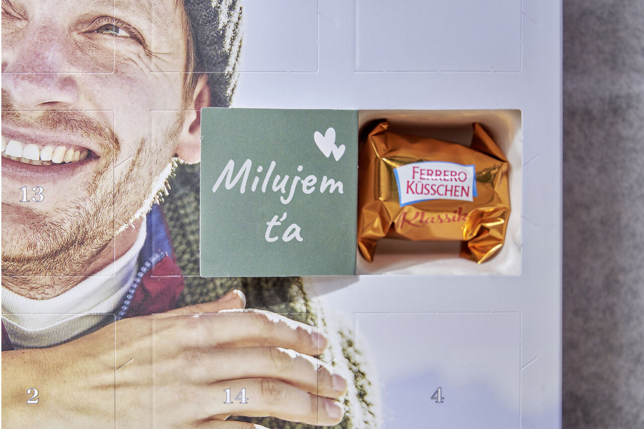 Na vnútornej strane otvoreného okna adventného kalendára je napísané „Milujem ťa“. Vedľa neho sú dve malé srdcia. V okne je Ferrero Küsschen.